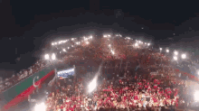 a large crowd of people are gathered in a stadium at night with a green and red flag behind them