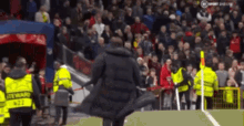 a man in a black coat is walking on a soccer field in front of a crowd of people .