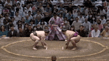 a sumo wrestler is kneeling down in front of a crowd while another wrestler stands in front of the referee