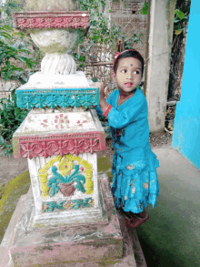 a little girl in a blue dress stands next to a colorful statue