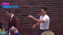 a man in a white shirt stands in front of a brick wall with a show de chistes sign above him