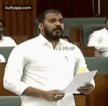 a man in a white shirt is giving a speech in a parliament while holding a piece of paper in his hand .