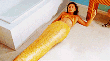 a woman in a mermaid costume is laying on the floor in front of a bathtub