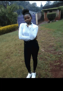 a woman in a white top and black pants stands in a grassy field