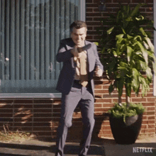 a man in a suit is holding a gun in front of a brick building and a potted plant .