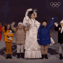 a woman in a white dress stands in front of a group of children with the olympic rings in the background