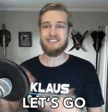 a man with a beard wearing a klaus let 's go shirt stands in front of a microphone