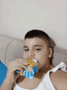 a young boy in a white tank top is eating a bag of chips .