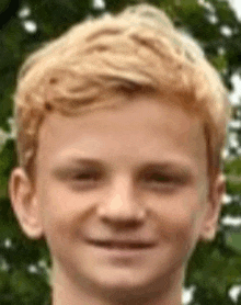 a young boy with blonde hair is smiling for the camera while standing in front of trees .