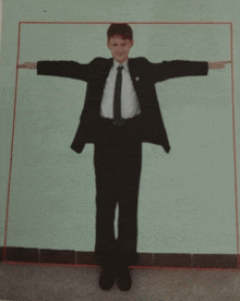 a boy in a suit and tie stands in front of a green wall with his arms outstretched