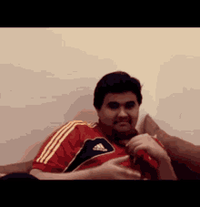 a man wearing a red adidas shirt sits on a bed