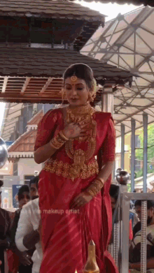 a woman wearing a red saree and gold jewelry is standing in front of a crowd and a sign that says makeup artist on it