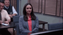 a woman in a suit and red shirt is sitting at a desk in front of a computer in a classroom .