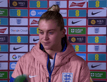 a woman in a pink jacket stands in front of a wall of advertisements including budweiser and m & s