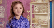 a man with long hair wearing a blue shirt is standing in front of a shelf filled with crowns .