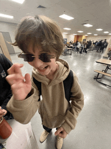 a young boy wearing sunglasses and a tan hoodie is standing in a cafeteria