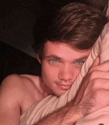 a shirtless young man with blue eyes is laying in bed with a pillow covering his face .