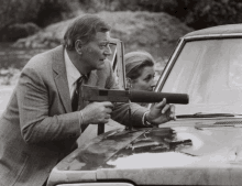 a man in a suit holds a gun while a woman looks on