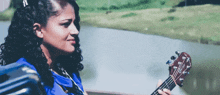 a woman with curly hair is holding a guitar in front of a lake