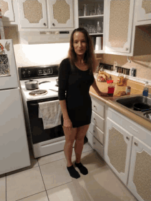 a woman in a black dress stands in a kitchen near a stove