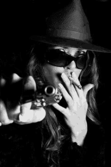 a woman wearing a hat and sunglasses is smoking a cigarette and pointing a gun at the camera .