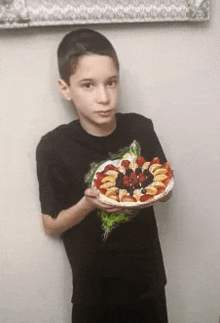 a young boy in a black shirt holds up a plate of fruit