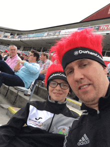 a man wearing an adidas jacket sits next to a boy wearing a red hat that says durham cricket