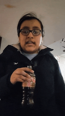 a girl wearing glasses and a hollister jacket is holding a coca cola bottle