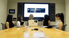 a group of women sit around a conference table looking at a large screen that says 16:21 on the wall behind them