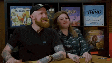 a man and a woman are sitting in front of a shelf full of board games including meadow and the thing