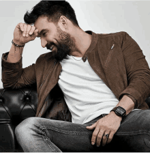 a man with a beard wearing a watch and a ring sits on a couch