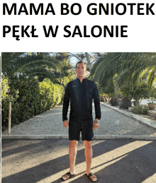 a man in a black jacket and shorts is standing in front of a sign that says mama bo gniotek pekl w salonie