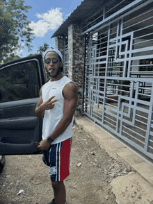 a man wearing a white tank top and red shorts is standing in front of a car