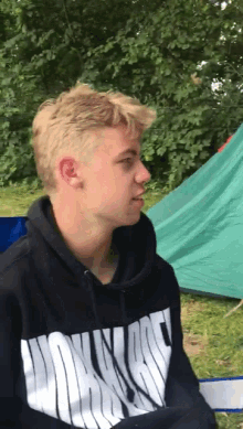 a young man wearing a black hoodie with a white lettering on the front is sitting in front of a tent