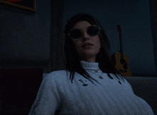 a woman wearing sunglasses and a white sweater sits on a couch in front of a guitar