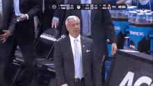 a man in a suit and tie stands in front of a ncaa scoreboard