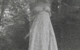 a black and white photo of a woman in a wedding dress standing in front of trees .