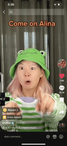 a woman wearing a green hat and a green and white striped shirt is pointing at the camera and says come on alina