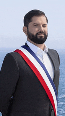 a man in a suit with a red white and blue sash around his neck stands in front of the ocean .