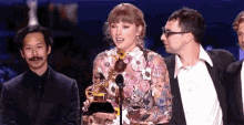 a woman in a floral dress is holding a trophy on a stage while standing next to two men in suits .