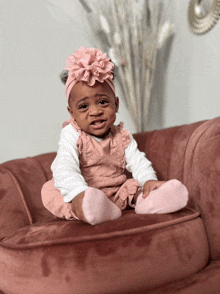 a baby girl wearing a pink headband and overalls sits on a pink couch