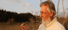 a man with a beard and glasses is standing in a field looking at a plant .