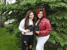 two women standing next to each other in front of a tree holding a book that says ' bible ' on it