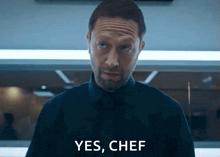 a man in a black shirt and tie is standing in a kitchen and saying yes , chef .
