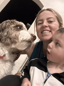 a woman is holding a child and a dog and the dog is licking the child 's face