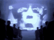 a group of people are standing in front of a large screen with a man 's face projected on it .