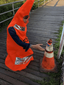 a person in an orange cone costume reaches for a traffic cone on a wooden walkway