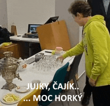 a man in a yellow hoodie is standing next to a table with glasses and a samovar .