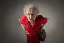 an elderly woman in a red dress and glasses is holding a briefcase and looking at the camera .