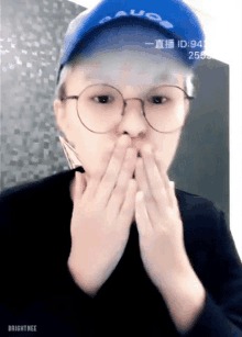 a person wearing glasses and a blue hat is covering their mouth with their hands .